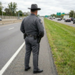 Highway patrol officer standing on shoulder of road facing traffic